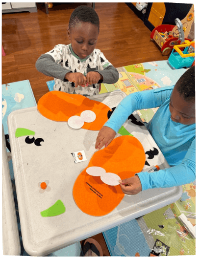 Two boys focused on a creative pumpkin craft project at a table indoors, an activity at Mama Bear Daycare's daycare program.