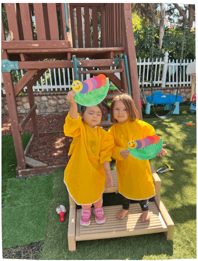 Two toddlers in yellow smocks holding craft projects outdoors, happy in the daycare program at Mama Bear Daycare.