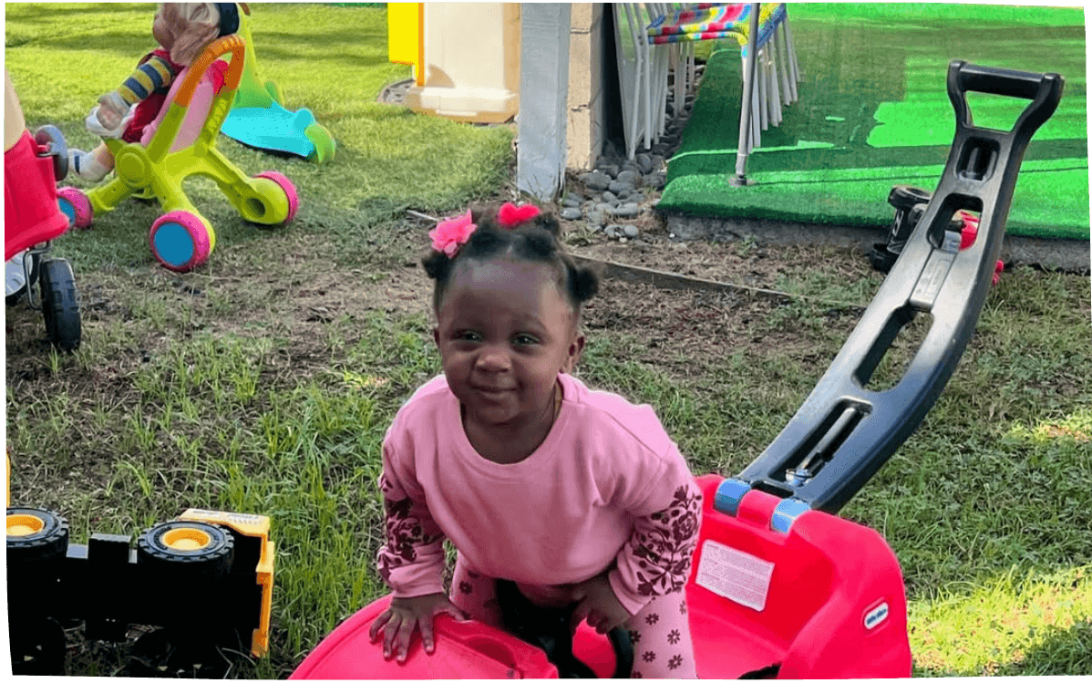 Toddler girl playing on a red ride-on toy outside on grass, enjoying outdoor time in Mama Bear Daycare's daycare program.